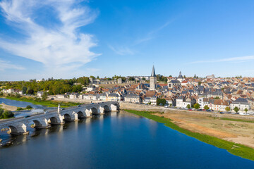 Obraz premium Wide view of La Charite sur Loire featuring a historic stone bridge crossing the Loire river, traditional rooftops, and a prominent church spire under a clear blue sky. The image highlights the blend