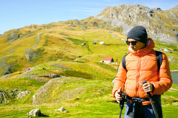 Male traveler with trekking poles walking through grassy mountain plateau near Kapetanovo Lake area. Symbolizes active lifestyle choice, hiking culture growth, and connection with environment.
