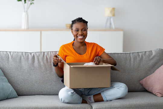 Full length of joyful black lady opening carton package, sitting on sofa with crossed legs, smiling at camera indoors. Young female consumer unboxing online store order, happy with delivery service