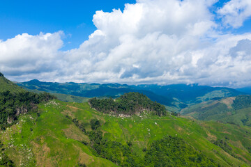 Obraz premium Wide aerial view of rolling green hills and forested ridges beneath a sky filled with large, textured clouds in the highlands between Phou Khoun and Luang Prabang, Laos. The landscape features vibrant