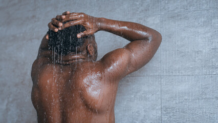 A man stands in a shower with his hands on his head. Water cascades down from above. The bathroom...