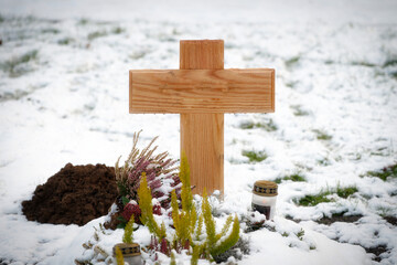 Close-up of a plain wooden cross on a snow-covered grave. Winter cemetery atmosphere with heather flowers and a memorial candle in Germany
