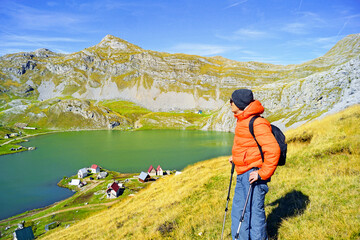 Hiker in orange jacket standing on alpine slope near Kapetanovo jezero, Montenegro. Represents outdoor adventure mindset, solo mountain exploration, slow travel inspiration, and nature immersion trend