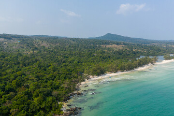 Naklejka premium Aerial perspective reveals dense green forest meeting the white sandy curve of Long Set Beach on Koh Rong island, Cambodia. Clear turquoise water, gentle waves, and rocky outcrops create a tranquil