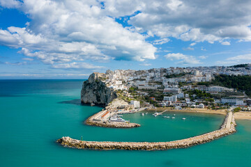Aerial perspective of Peschici's marina with curving stone breakwaters, turquoise water, and a striking hillside of whitewashed buildings atop dramatic cliffs beneath a sky filled with scattered