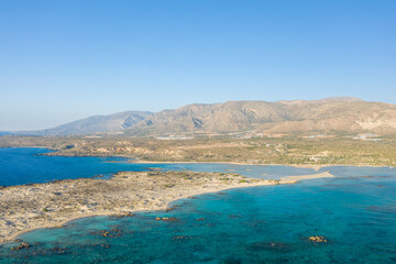Aerial view of a rugged coastal peninsula at Elafonisi beach in Crete, Greece, with vivid turquoise waters meeting arid hills under a clear blue sky. The landscape is sunlit, expansive, and naturally