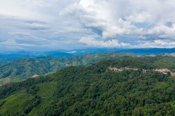 Naklejka premium A small rural settlement is nestled among densely forested hills, with expansive mountain ridges stretching into the distance under a sky filled with dramatic clouds. The scene captures the remote