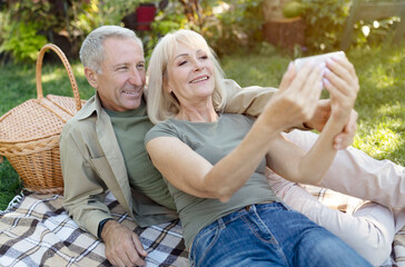 Fototapeta premium Happy senior spouses video calling on smartphone while having picnic and resting in garden at warm spring day, copy space. Elderly couple capturing happy moments, taking selfie