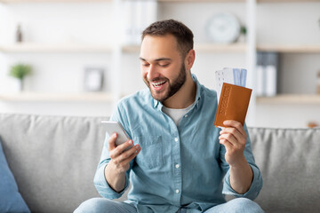 Happy young man holding international passport and plane tickets, using smartphone to book vacation...