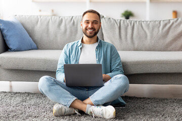 Full length of happy young guy sitting cross legged on floor, using laptop computer, smiling at...