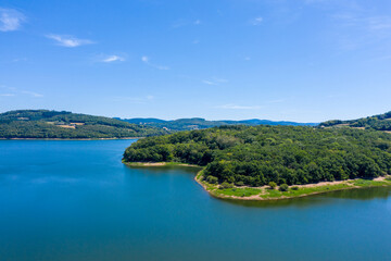 Fototapeta premium Aerial view of a lush green peninsula extending into the vivid blue waters of Panneciere-Chaumard reservoir, surrounded by rolling wooded hills and open farmland in Burgundy. The clear summer sky and