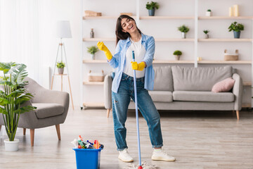 Happy young woman cleaning her flat, singing and listening to music. Excited maid dancing and holding mop, having fun, free space. Millennial housewife in protective gloves enjoying domestic chores