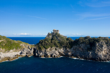 Fototapeta premium A historic stone watchtower crowns the rugged Capo di Conca promontory in Italy, surrounded by lush Mediterranean vegetation and overlooking deep blue sea. The dramatic coastline is set beneath a