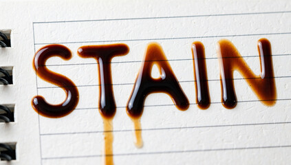 Close-up of "Stain" Text Written in Dark Brown Liquid on Notebook Paper