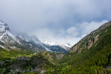 Fototapeta premium Snow-capped mountains rise above lush green valleys and scattered alpine villages in Val d'Aoste, Italy. The landscape features dramatic contrasts, forested slopes, and a mix of sunlight and drifting