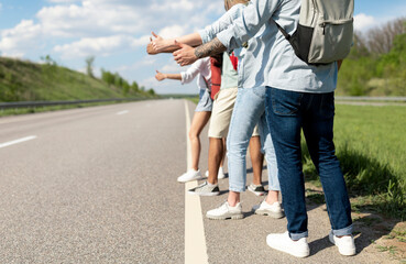 Fototapeta premium Group of unrecognizable young friends standing along road, making thumb up gesture, hitchhiking for car during road trip, traveling together in summer, closeup. Outdoor vacation, tourism concept