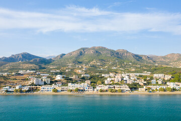 Panoramic view of a seaside village in Analipsi, Crete, with whitewashed buildings lining a sandy beach and lush green hills rising in the background under a clear blue sky.
