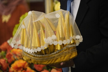 A close-up shot of a man in a black suit carrying a traditional Indonesian wedding gift (seserahan). The gift consists of a bunch of bananas individually wrapped in elegant gold cone-shaped paper, arr