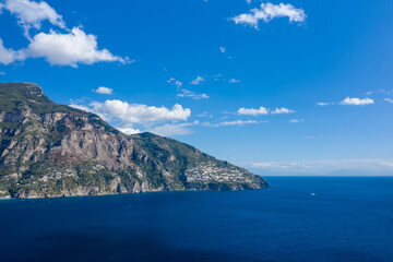 Obraz premium Steep rocky cliffs covered with green vegetation rise above deep blue Mediterranean waters beneath a vivid sky. Sunlight highlights the natural textures and crisp outlines of the Italian coastline