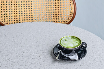 Close up view of hot matcha latte in the black mug or cup. spoon. stainless steel. tissue. table. grey or gray. white. green. copy space, empty, free, negative, text, design. wooden chair. rattan. gra