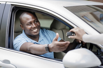 Cheerful black man taking key from car from dealer, renting auto for summer trip, side view. Happy african american guy buying new automobile, sitting inside the car and taking automatic key