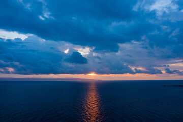 Vivid blue clouds gather above a calm sea as the sun sets on the horizon near the Laconia coastline, casting a striking reflection across the water and creating a moody, atmospheric scene.