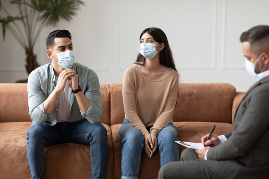 Marital Therapy. Male psychotherapist in face mask interviewing young family, listening and taking notes. Millennial arab couple sitting on couch, sad spouses having session with male psychologist
