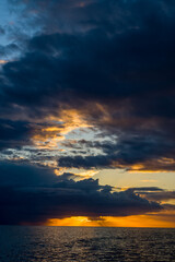 Deep blue and golden clouds gather above the Adriatic Sea at twilight, with dramatic bands of light breaking through the sky and reflecting on the calm water. The scene captures a striking interplay