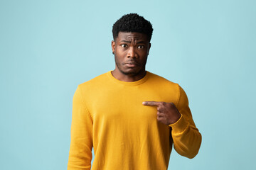 Confused bearded young black guy in yellow cardigan pointing at his chest, posing on blue studio...