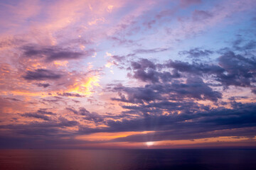 Fototapeta premium Golden and lavender clouds scatter across the sky as the sun sets above the calm Mediterranean Sea, casting soft reflections and creating a luminous, tranquil atmosphere.
