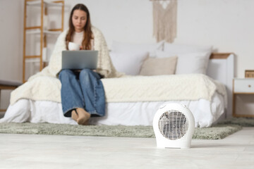 Young woman sitting on bed with laptop and cup of tea near electric fan heater in bedroom