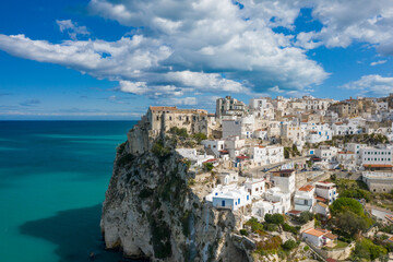 Sweeping view of Peschici's whitewashed houses clustered atop dramatic limestone cliffs above the vivid turquoise Adriatic Sea, under a sky filled with dynamic clouds.