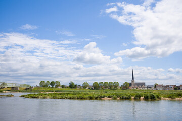 Obraz premium Peaceful riverside scene with a pointed church steeple, leafy trees, and a metal bridge spanning the Loire under a bright blue sky with scattered clouds.