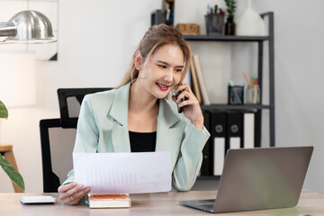 Smiling Asian businesswoman talking on a smartphone while reviewing financial documents at her desk...