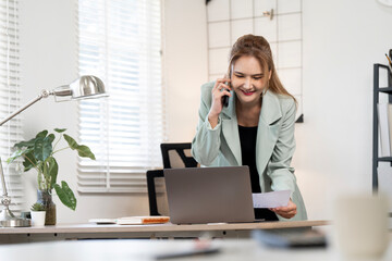 Cheerful Asian businesswoman discussing paperwork on a mobile call while looking at her laptop...