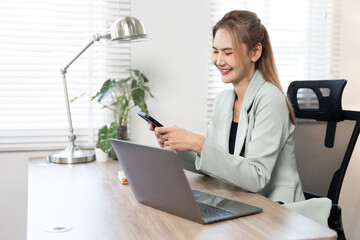 A cheerful Asian businesswoman smiling while checking her smartphone and working on a laptop at a...