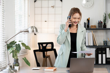 Happy Asian businesswoman holding document and talking on smartphone while standing at office desk,...