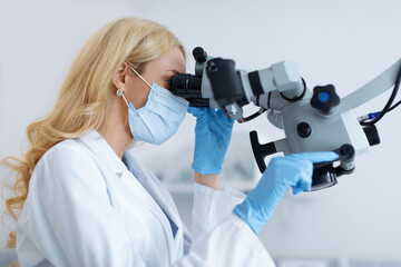 Side view of blonde woman stomatologist in workwear looking at modern dental microscope, using...