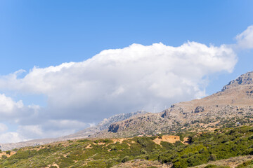 Sparse wind turbines dot a rugged mountain slope in Crete, with scattered green shrubs and dramatic clouds drifting across a bright blue sky.