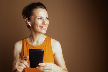 A woman in a yellow tank top and white wireless earbuds smiles and points at her smartphone, conveying casual tech use and communication.