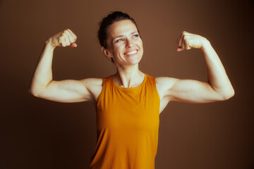 An empowered woman in a mustard-yellow tank top flexes her biceps with raised, clenched fists and a broad smile, embodying strength, confidence, and well-being.
