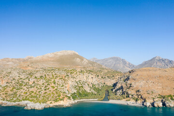 Expansive aerial view of Preveli beach in Crete, featuring rugged rocky cliffs, dry hills, and a narrow river meeting the clear blue sea under a cloudless sky. The landscape is defined by contrasting