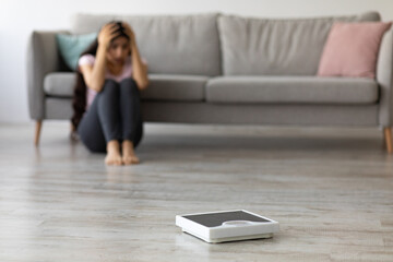 Stressed young Indian woman sitting on floor, grabbing her head in terror, looking at empty scales,...