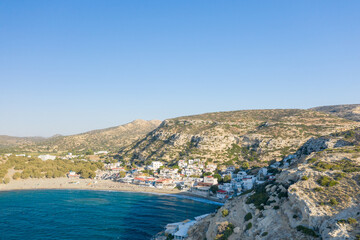 Fototapeta premium Matala village sits nestled between a sandy bay and sunlit limestone hills on the southern coast of Crete. The clear blue water contrasts with the textured rock formations and whitewashed buildings