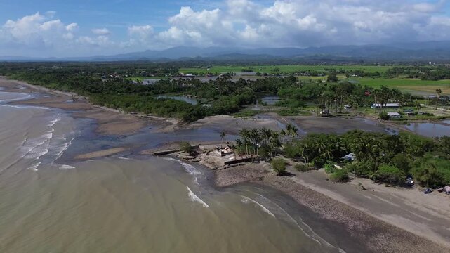 Aerial drone footage of a river mouth in Roxas Oriental Mindoro where freshwater flows into the sea, surrounded by sandy shoreline, palm trees, farmland and distant mountains.