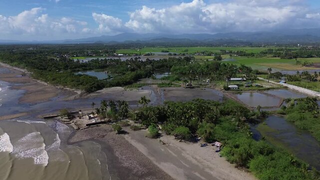 Aerial drone footage of a river mouth in Roxas Oriental Mindoro where freshwater flows into the sea, surrounded by sandy shoreline, palm trees, farmland and distant mountains.