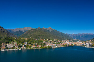 Fototapeta premium Expansive panorama of Omegna on the northern shore of Lake Orta in Italy, with modern waterfront buildings, lush green hills, and distant mountain peaks beneath a vivid blue sky. The calm lake water