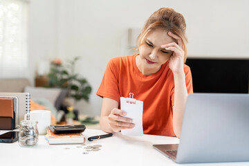 Stressed young Asian woman looking at a bill or receipt while working at home. Feeling worried...