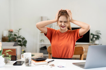 Overwhelmed young Asian woman holding her head in stress while working at a desk with a laptop and...