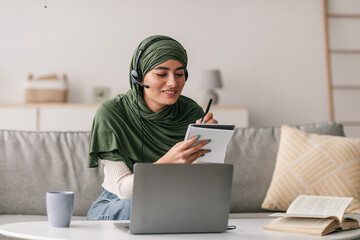 A cheerful young Arab woman in a hijab is engaged in an online lesson at home. She wears headphones and takes notes while using her laptop, showcasing the remote learning experience.